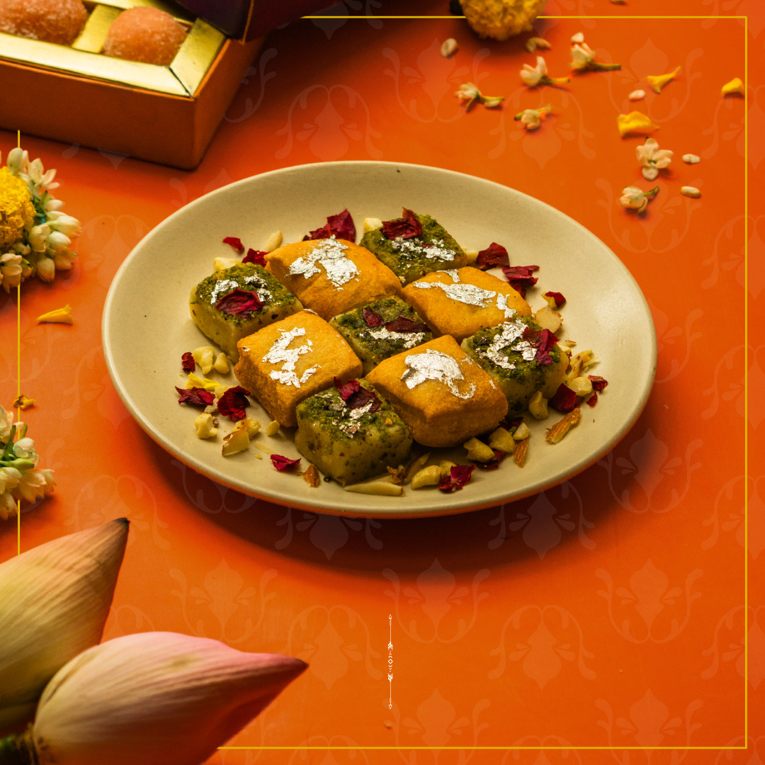 Plate of assorted Indian sweets garnished with silver leaf, rose petals, and chopped nuts, placed on an orange festive background with lotus flowers and marigolds.